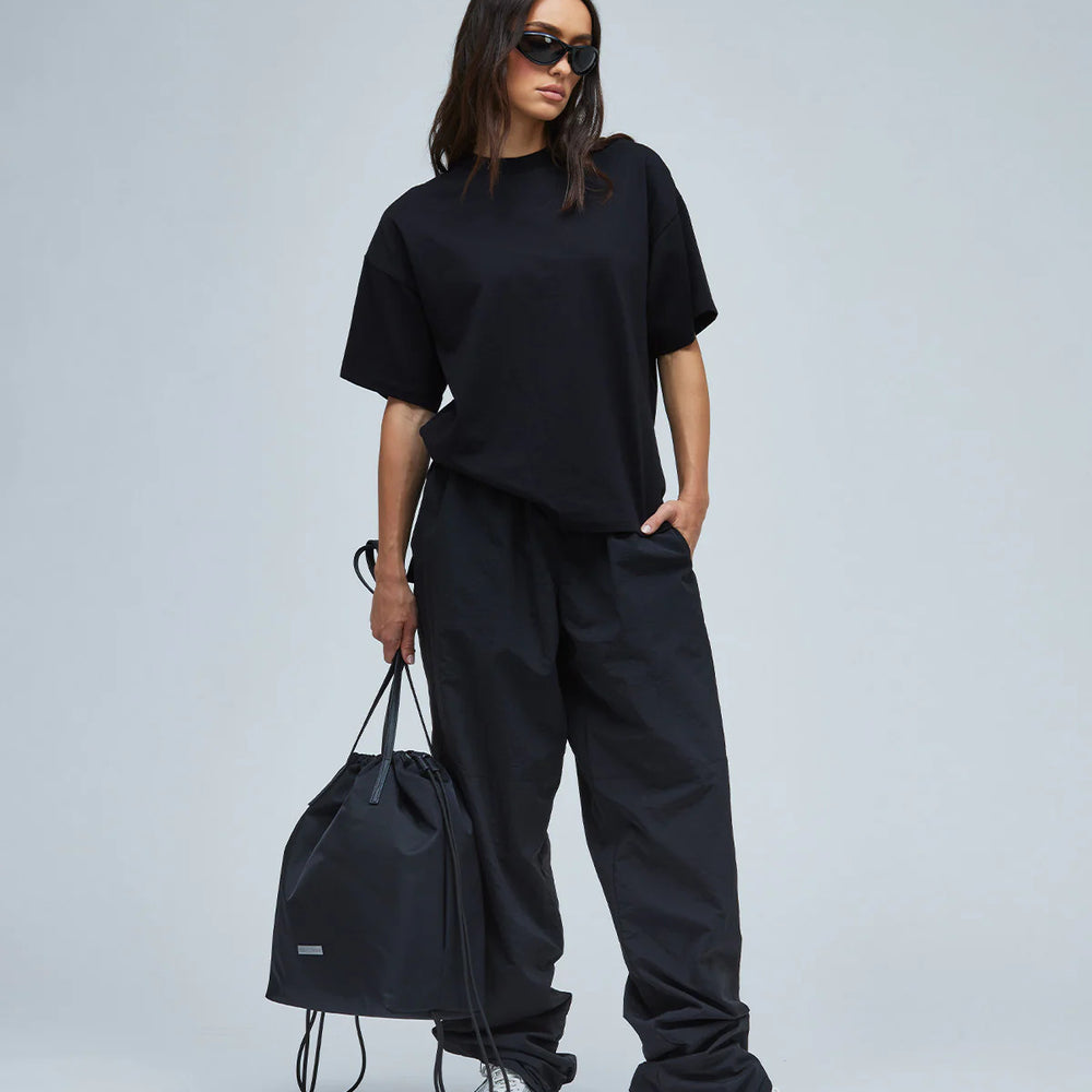 
                  
                    A model wearing black-colored women's nylon track pants & a T-shirt while holding a black bag
                  
                